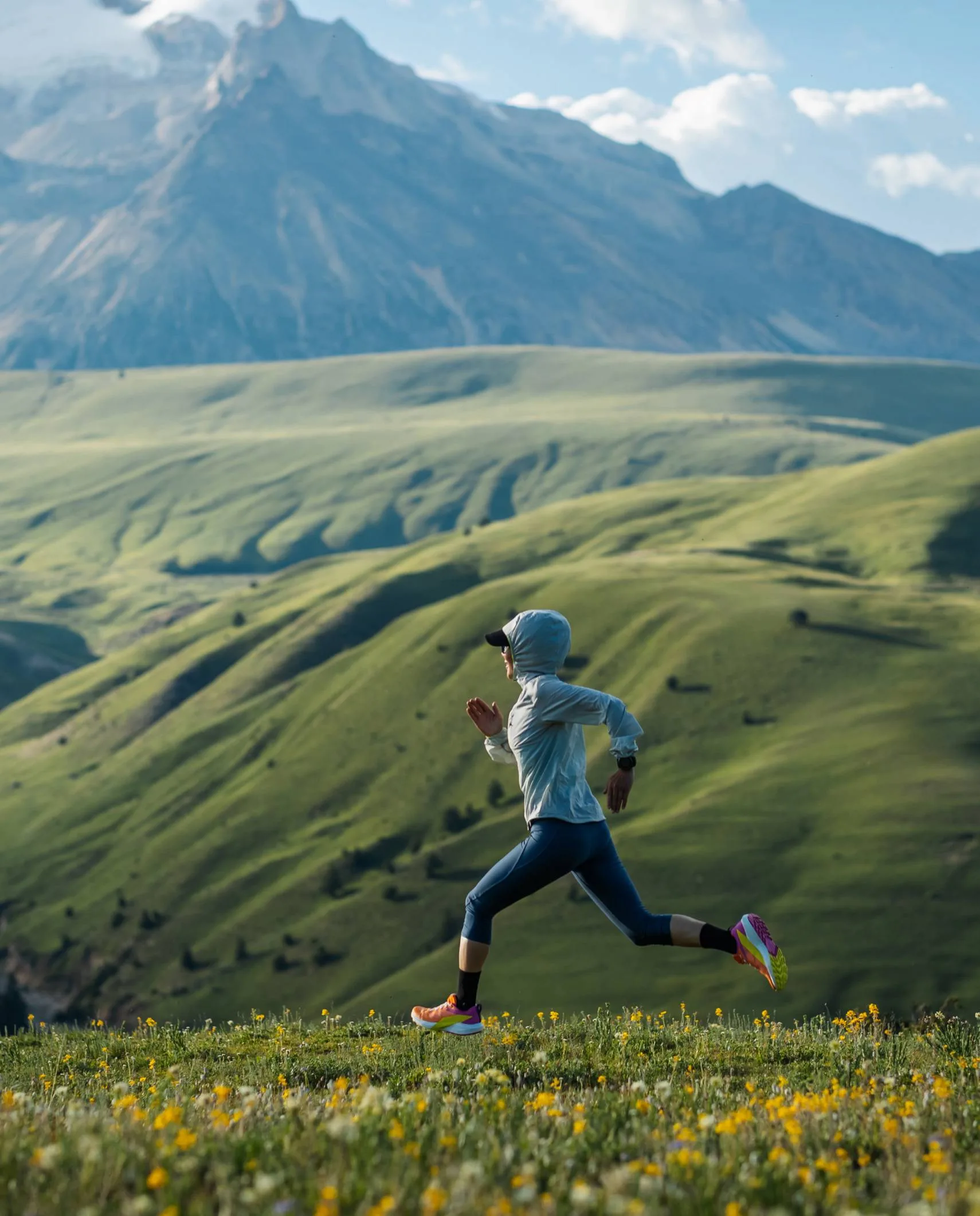 Trail runner crossing an alpine meadow beneath mountain peaks.